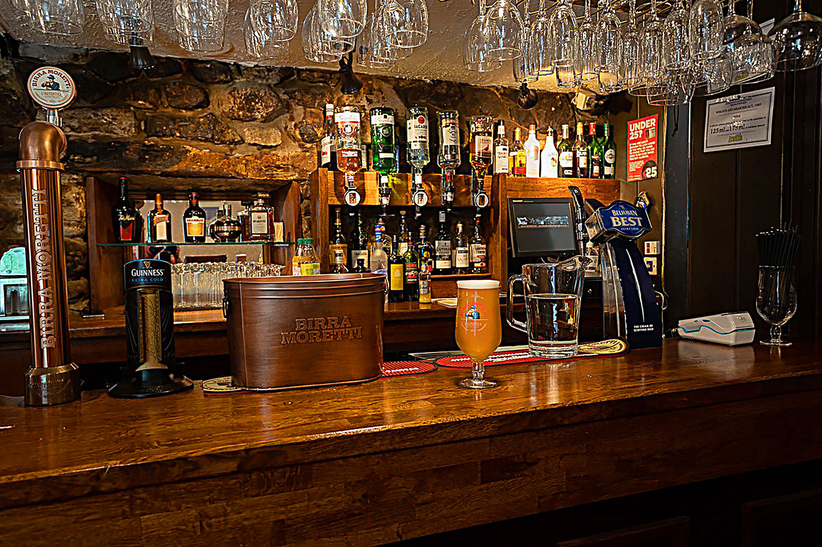 The bar at The Wheatsheaf Inn, with hanging glassware, stone walls and a pint on the counter.